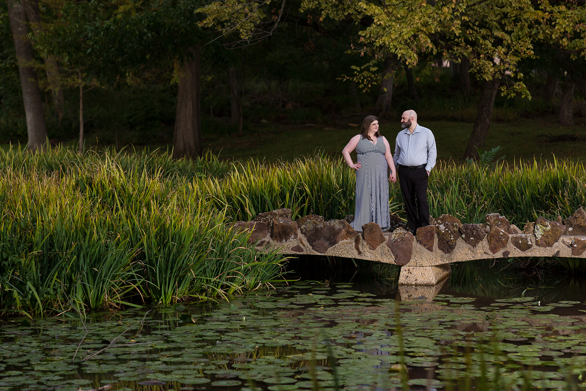 Dallas wedding photographer Stefani Ciotti Photography captures engagement photo at Texas Women's University with engaged couple standing together looking at each other on a bridge over a lake filled with Lilly pads with trees surrounding them