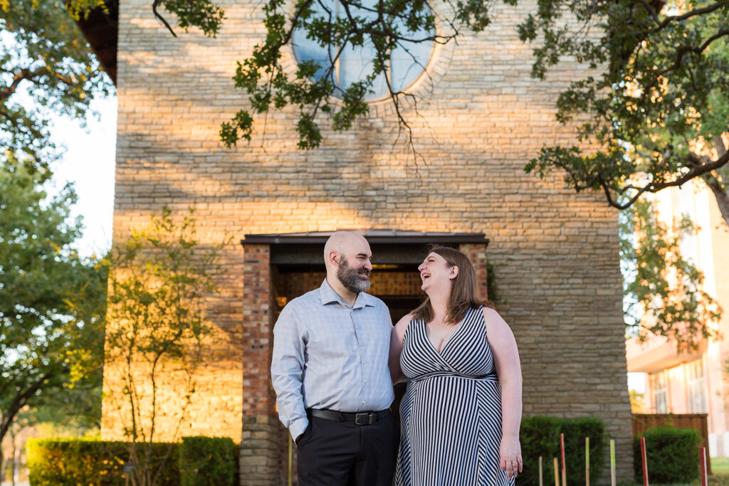 Dallas engagement picture of a man and woman in grey outfits laughing at each other and standing in front of an old building with a round window at Texas Woman's University