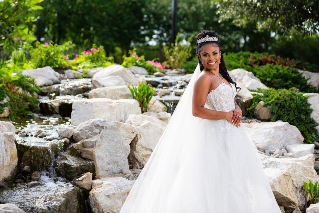 Bride wearing strapless wedding dress with a long veil and crown smiling in front of rock waterfall at Knotting Hill Place wedding venue in Dallas tx by Stefani Ciotti Photography