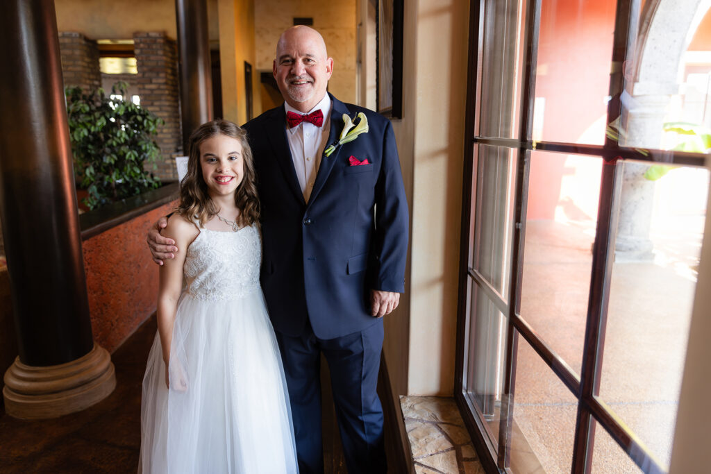Groom standing with granddaughter smiling by window in navy suit with red bowtie before Stoney Ridge Villa wedding ceremony