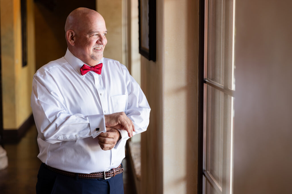 Stefani Ciotti Photography captures groom laughing by window while putting on cufflinks in navy suit with red bowtie before Stoney Ridge Villa wedding ceremony