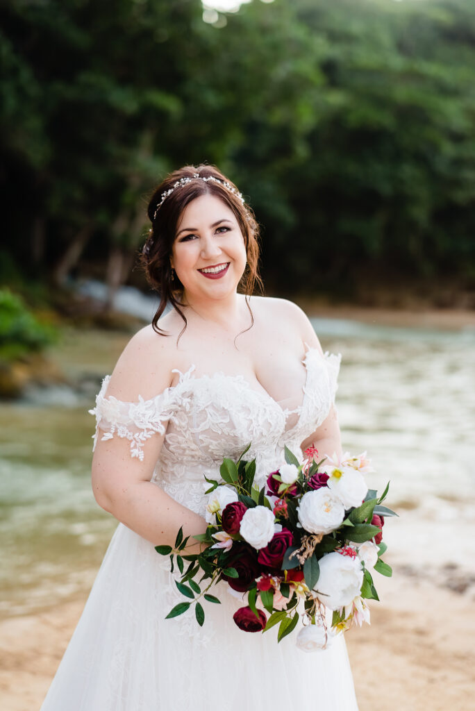 bride smiling at camera with red and white bridal bouquet after wedding ceremony on the beach with trees, water and waterfall in the background