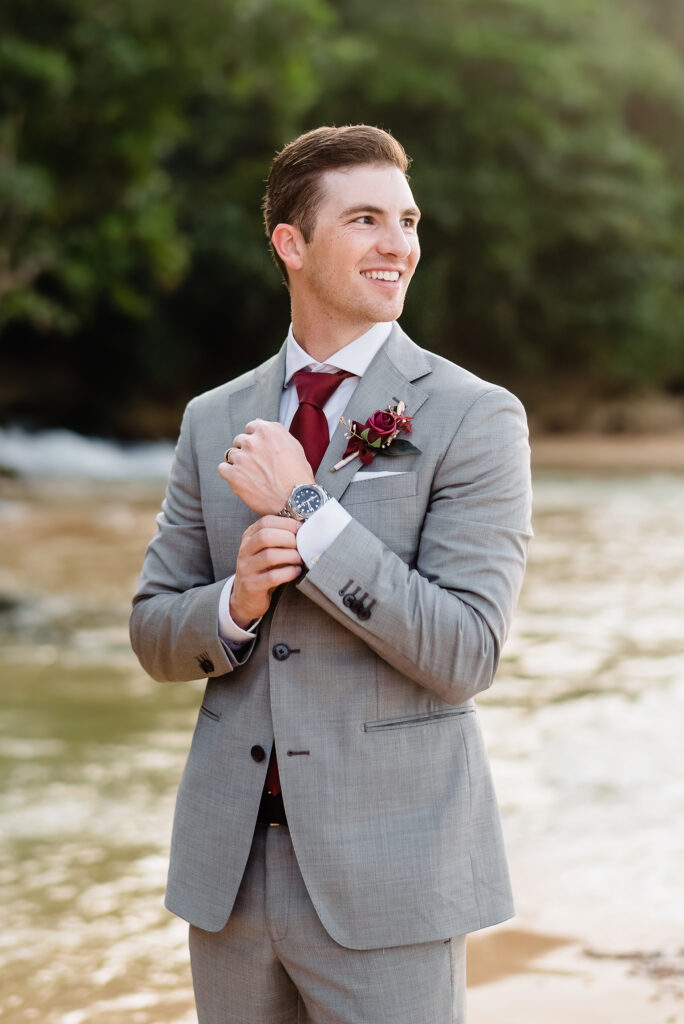groom smiling off camera adjusting cufflinks and watch after wedding ceremony on the beach with trees, water and waterfall in the background