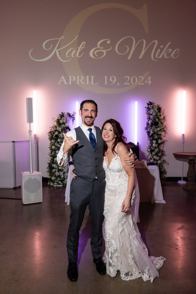 Bride and groom smiling on the dance floor with pink and purple uplighting with their custom monogram on the wall