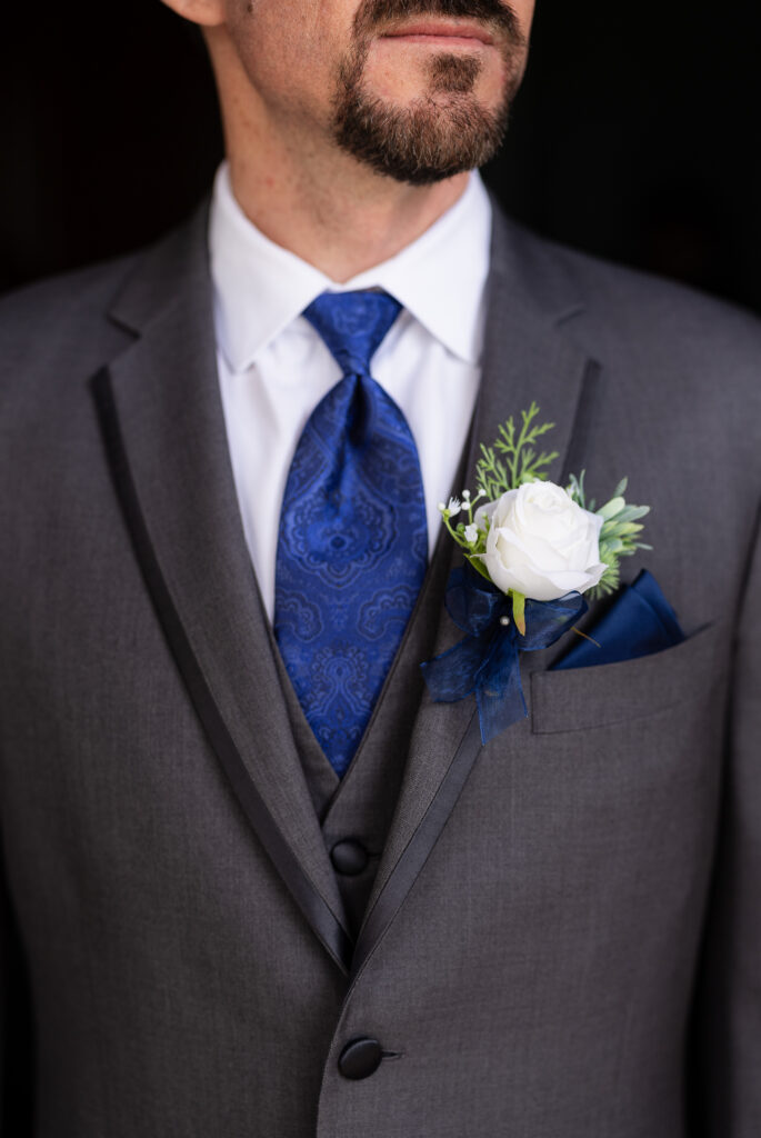 Close up of groom's grey suit, blue tie, blue pocket square and white flower boutonniere