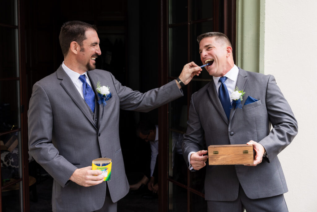 Groom gifting groomsmen a box of crayons and feeding him one as a gift as they both laugh