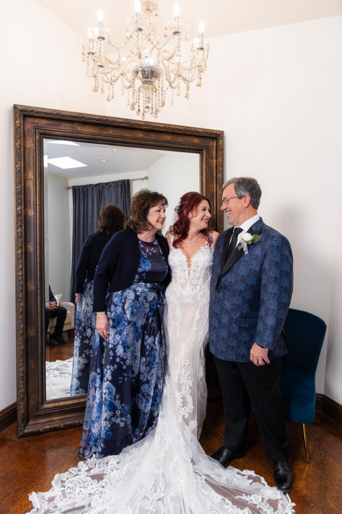 Bride hugging mom and dad laughing in front of large mirror in Stoney Ridge Villa bridal suite under crystal chandelier