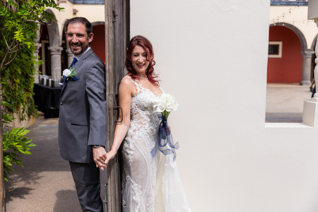 Bride and groom holding hands behind old wooden door for first touch while bride holds white tulip bouquet