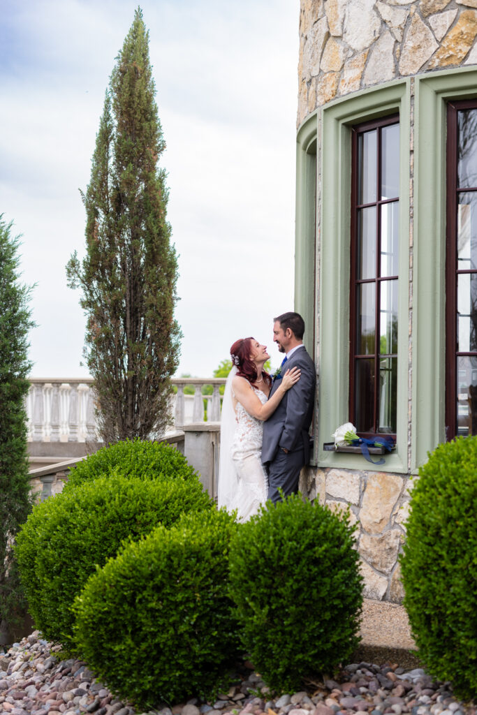 Bride and groom smiling at each other up against wedding venue surrounded by trees and bushes