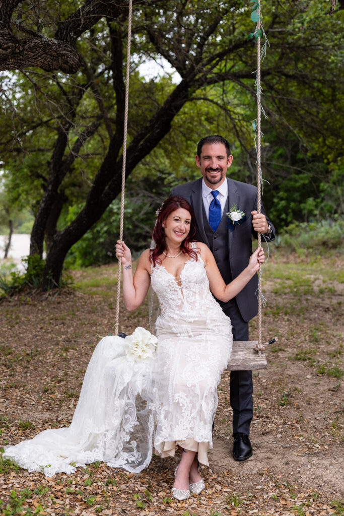 Bride sitting on tree swing with groom standing behind her smiling at camera