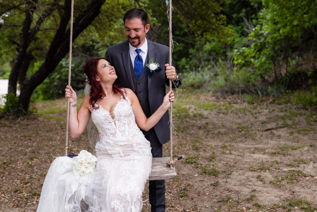 Bride sitting on tree swing while looking up at groom smiling down at her in grey suit