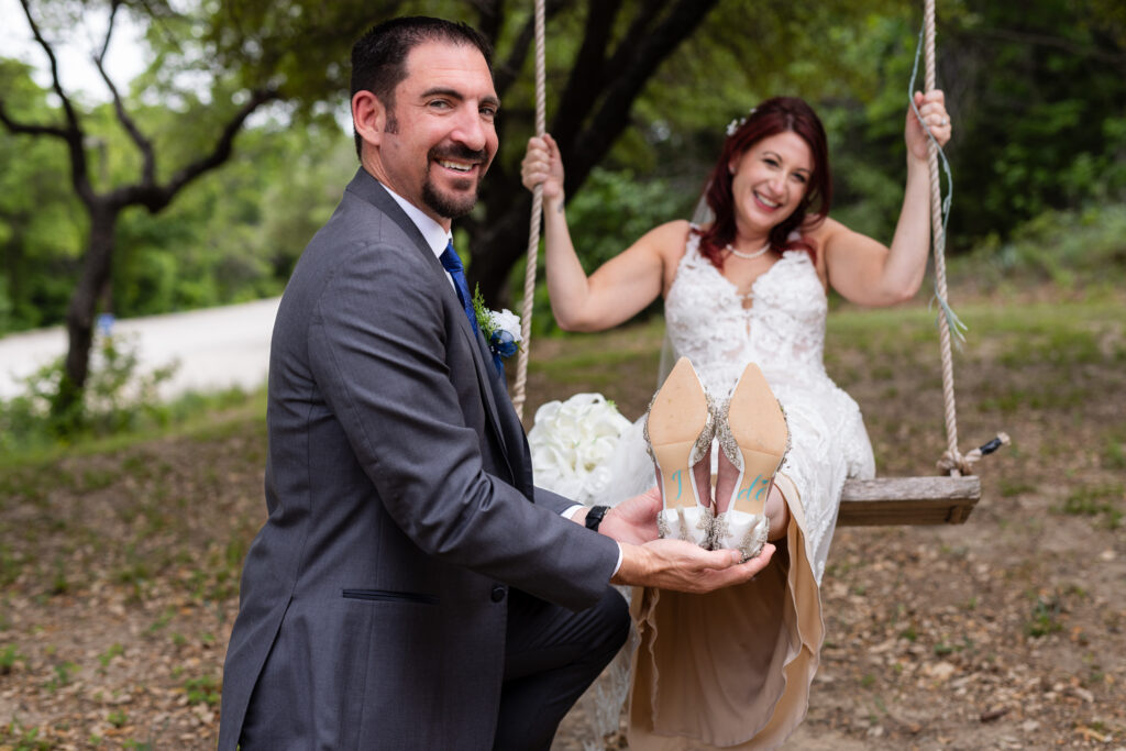 Bride sitting on tree swing with groom kneeling and holding up brides shoes that read "I DO"