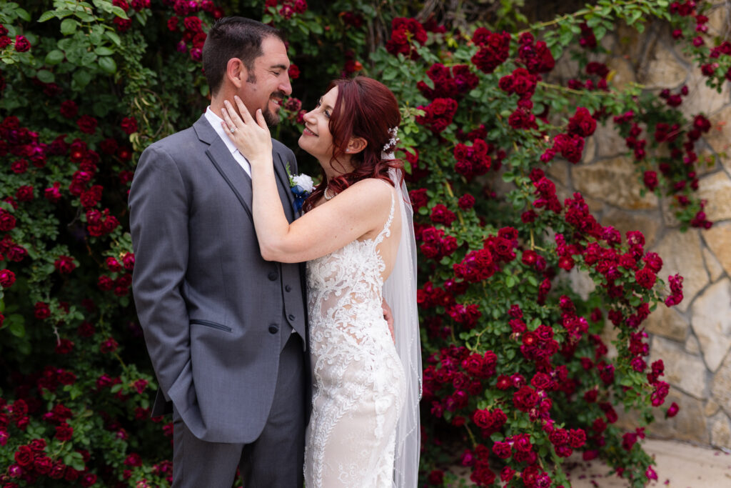 Bride and groom smiling at each other surrounded by red rose vines