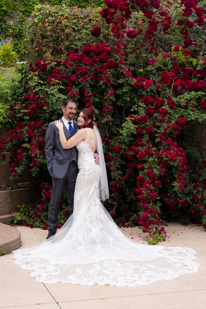 Red-haired bride smiling with one hand on groom's chest surrounded by red rose vines