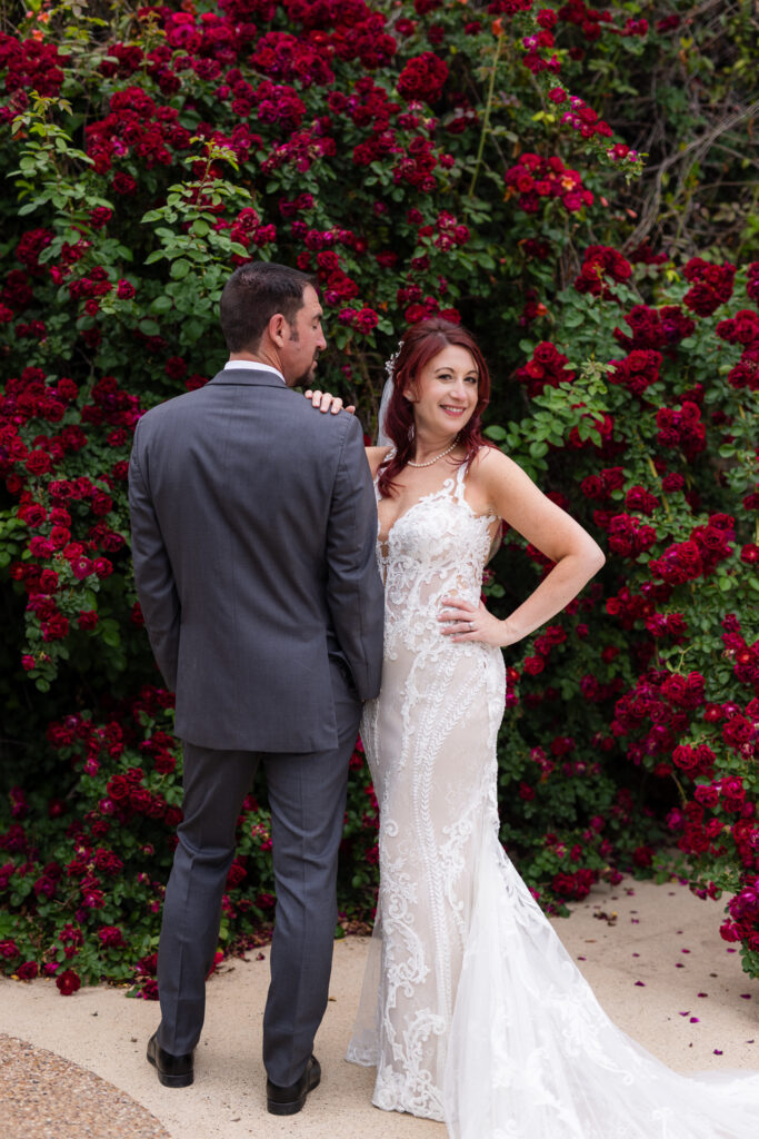 Red-haired bride smiling with one hand on hip and the other hand on her groom's shoulder while he looks over at her surrounded by red rose vines