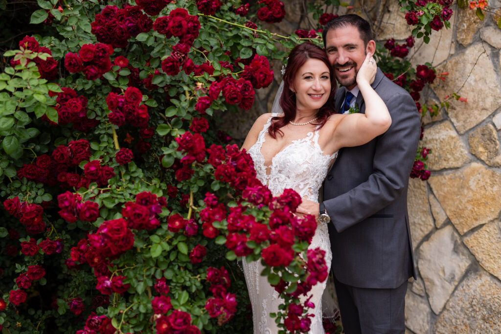 Bride posing with groom behind red roses at Azle TX wedding venue, Stoney Ridge Villa