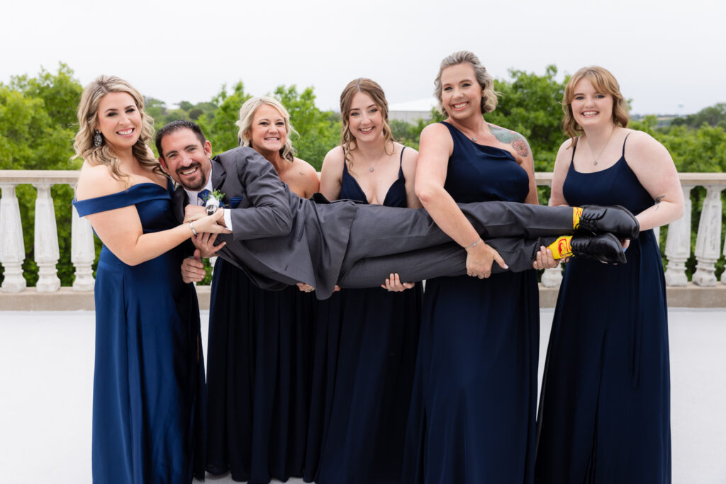 Bridesmaids in blue dresses holding groom in a grey suit laughing