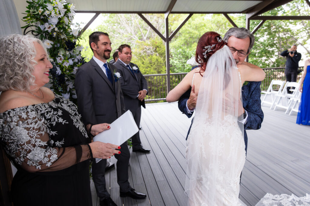 Dad hugging bride after walking her down the aisle while officiant and groom smile and look on