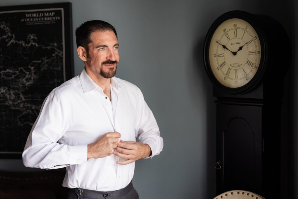 Groom buttoning up dress shirt with old clock in the background in groom's suite