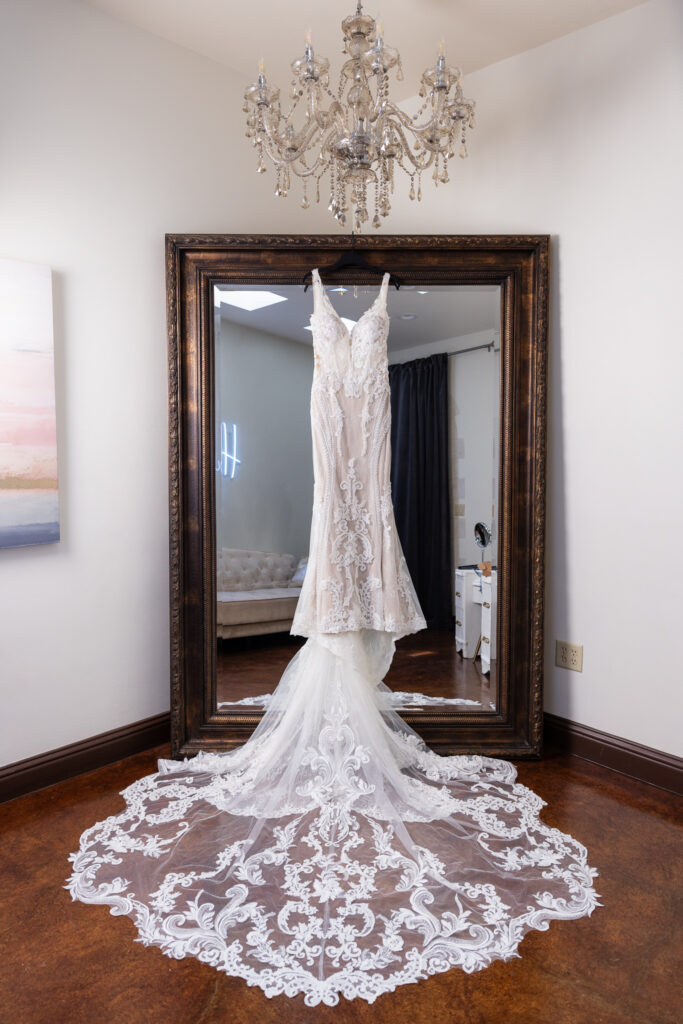 Wedding dress hanging on large mirror with long intricate and elegant train in bridal suite under crystal chandelier