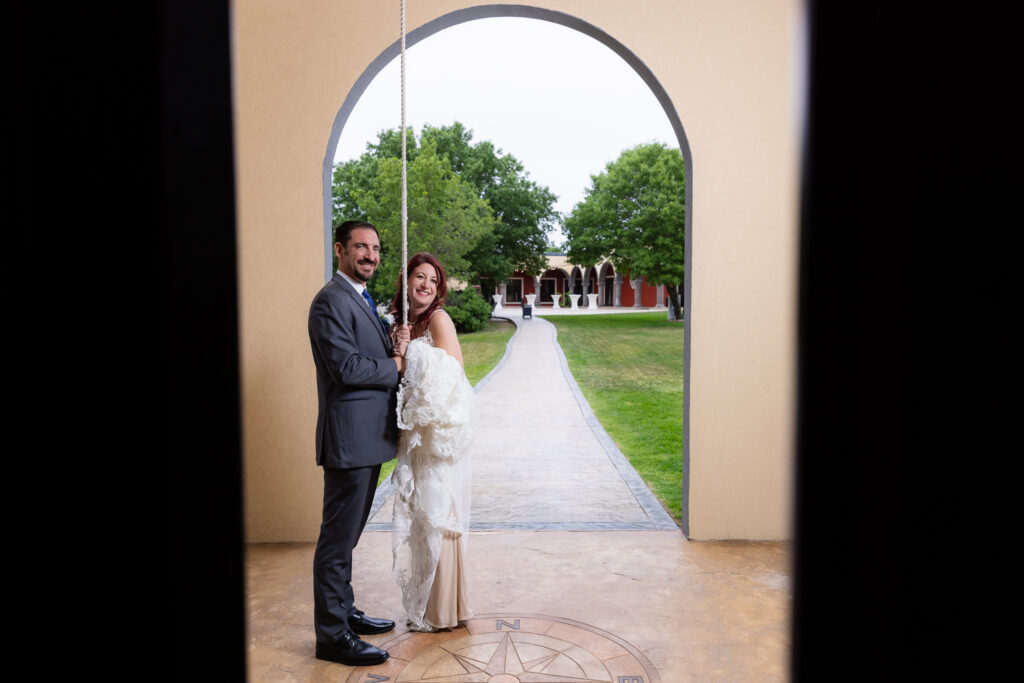 Bride and groom holding rope to bell tower smiling with Stoney Ridge Venue courtyard in the background