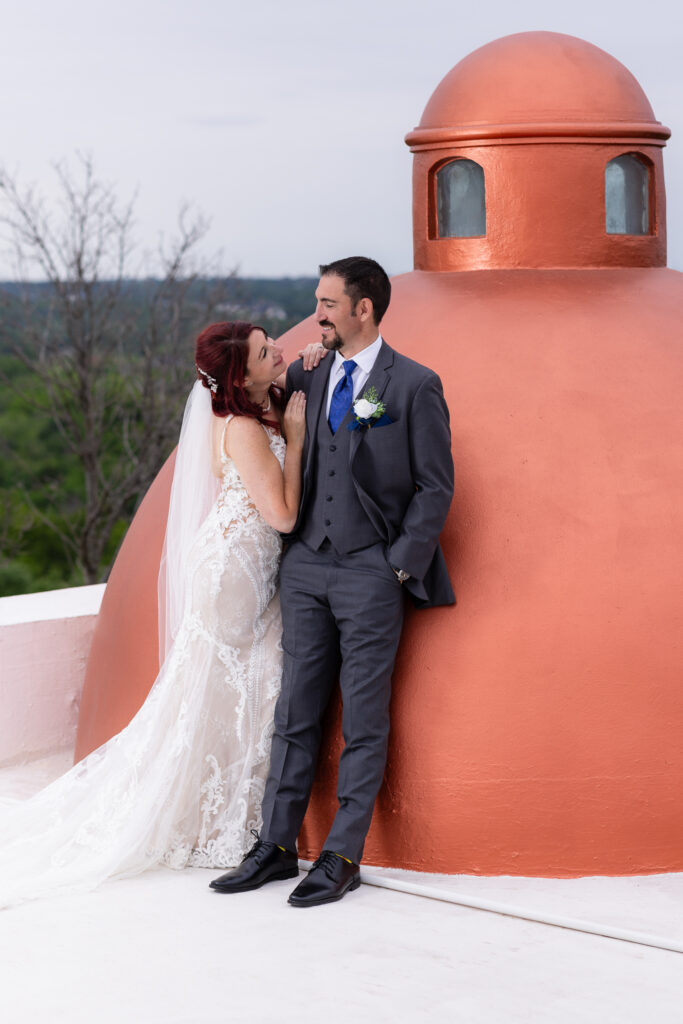 Red-haired bride intimately smiling at groom in grey suit laughs at her on rooftop