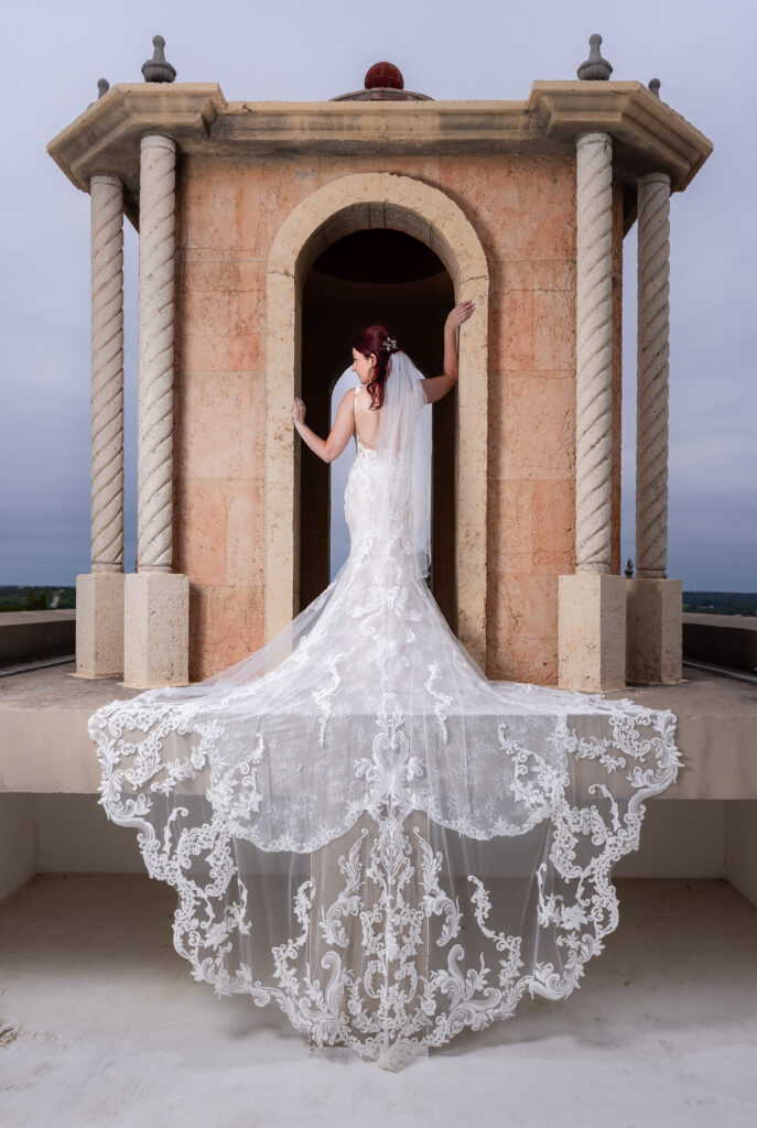 Red-haired bride posing on bell tower on Stoney Ridge Villa wedding venue's rooftop in Azle TX with intricate and laced wedding train flowing