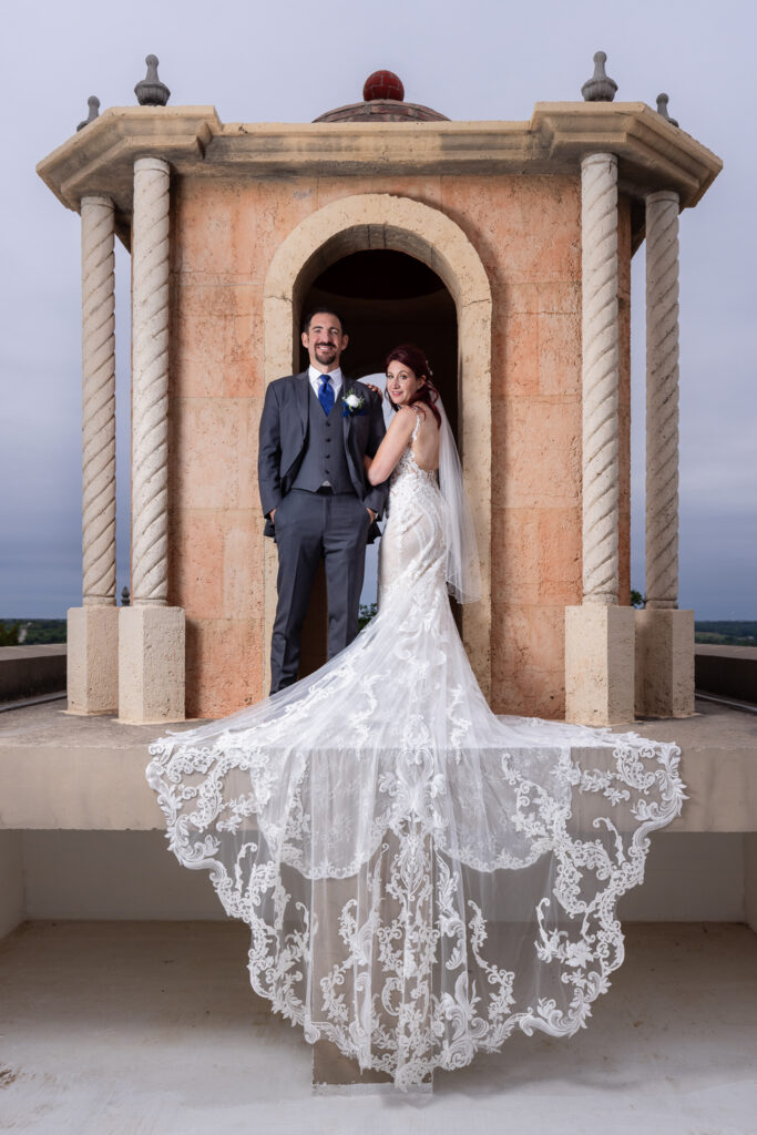 Bride and groom smiling at camera on bell tower on Stoney Ridge Villa wedding venue's rooftop in Azle TX with intricate and laced wedding train flowing