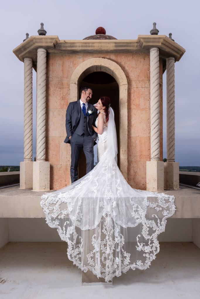 Bride and groom smiling at each other on bell tower on Stoney Ridge Villa wedding venue's rooftop in Azle TX with intricate and laced wedding train flowing