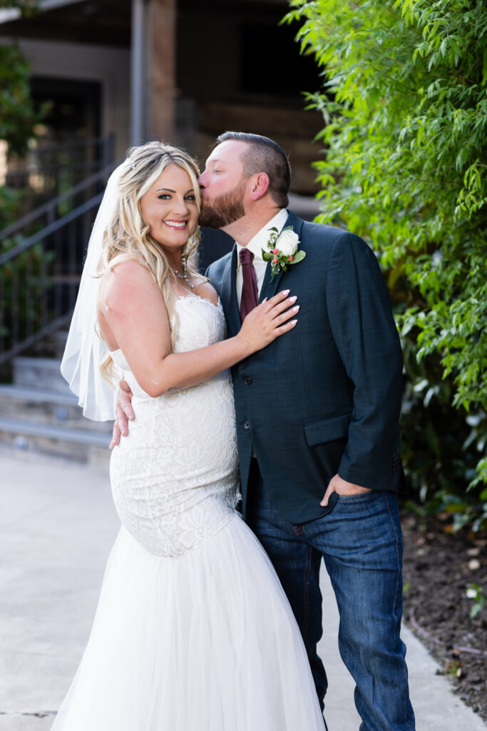 Groom kissing bride on temple while she smiles at the camera