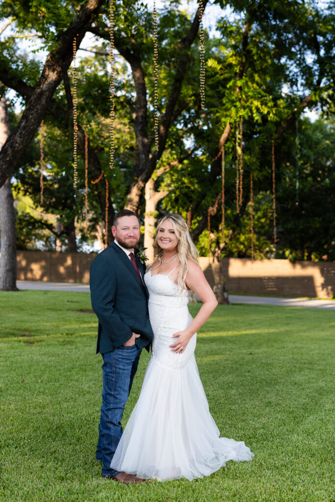 Wedding couple smiling under large lush trees with hanging string lights