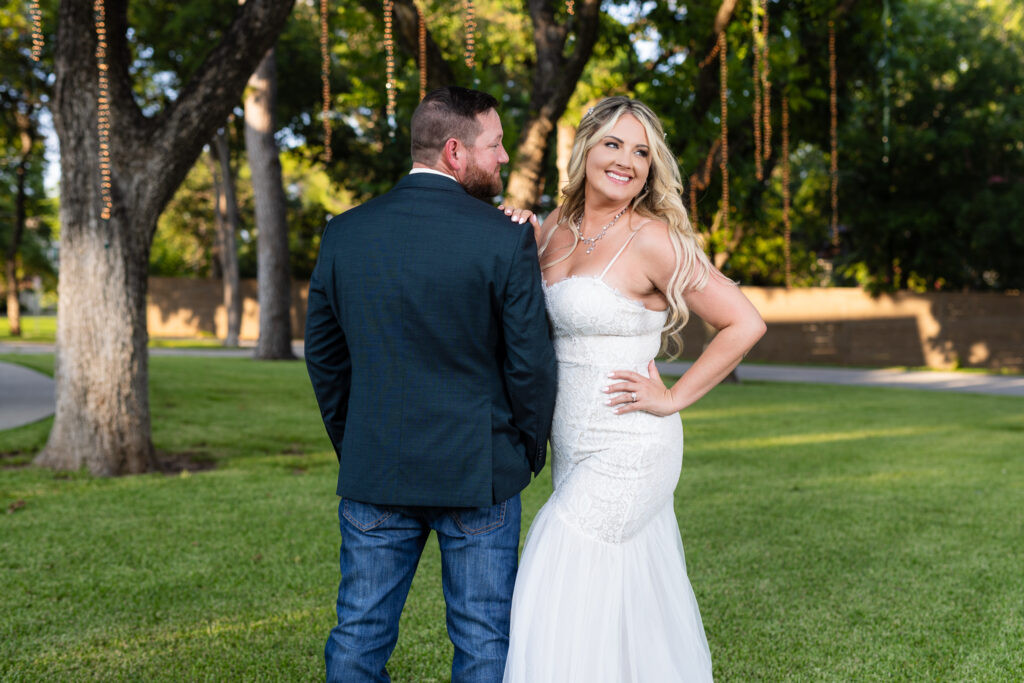 Bride smiling over shoulder while leaning on groom under large lush trees with hanging string lights