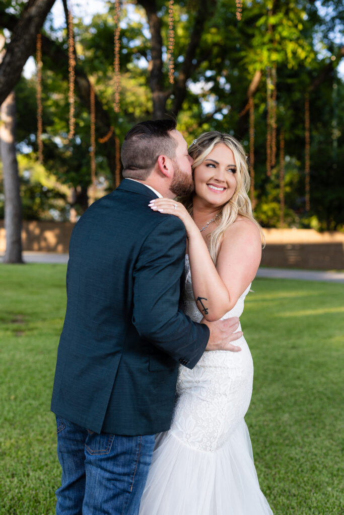 Groom kissing smiling bride on temple under trees and hanging string lights