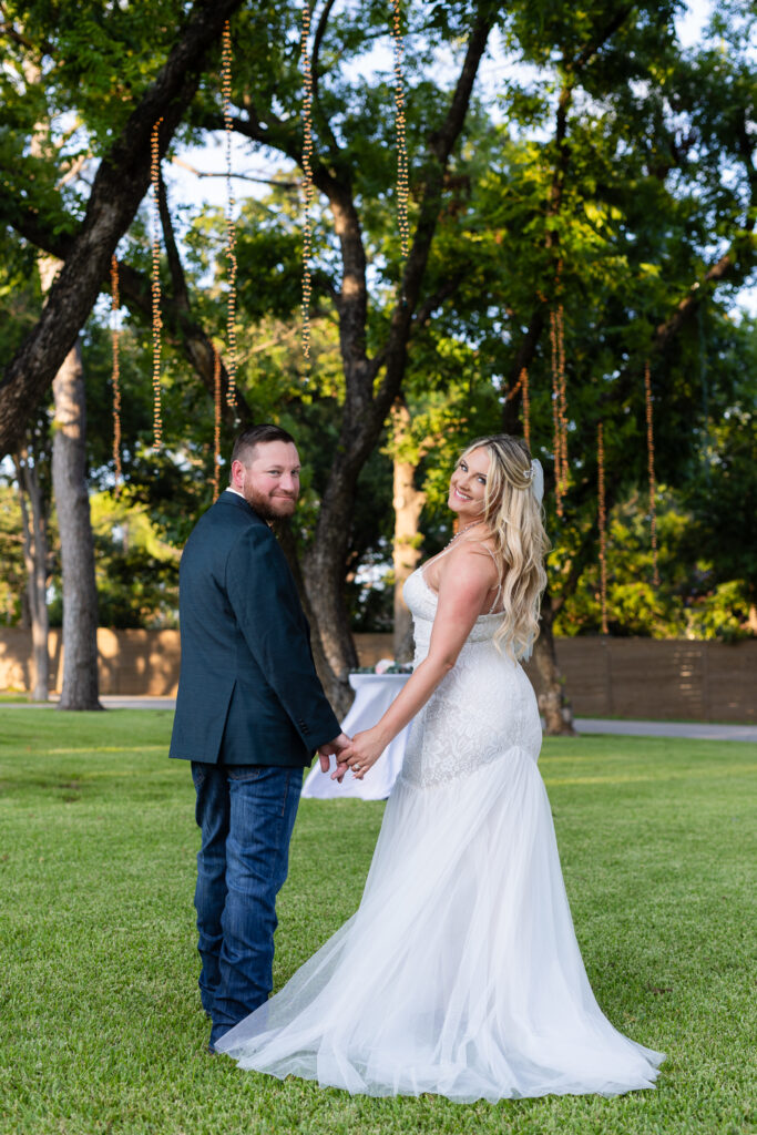 Groom holding brides hands while both looking back over their shoulders smiling under lush trees and hanging string lights after elopement ceremony at Hotel Lucy in Granbury TX