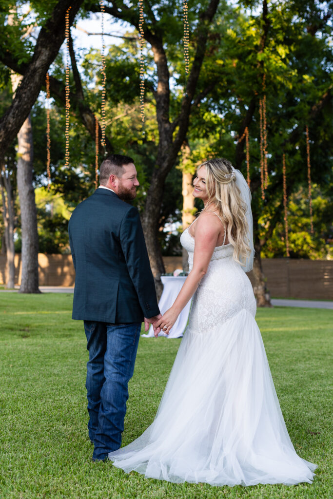 Groom holding brides hands while both looking back over their shoulders smiling at each other under lush trees and hanging string lights