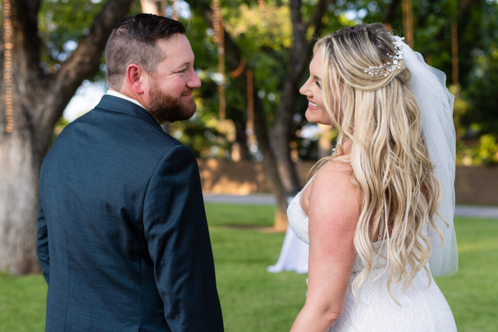 Groom holding brides hands while both looking back over their shoulders smiling at each other under lush trees and hanging string lights