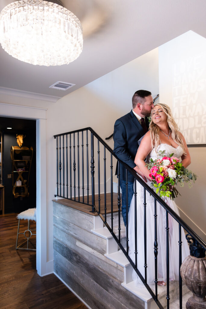 Bride and groom leaning against indoor staircase banister while groom kissing bride on forehead