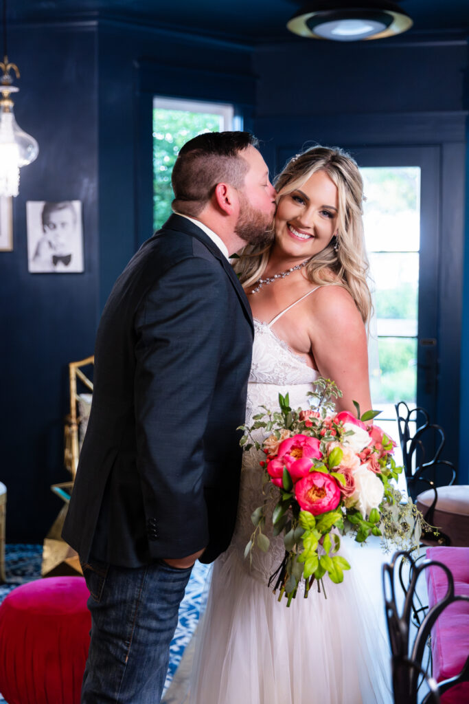 Bride holding pink flower wedding bouquet while groom kisses her cheek inside hotel bar