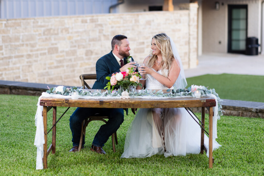 Bride and groom toasting at rustic sweetheart table smiling at each other outdoors