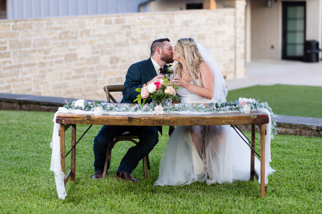 Bride and groom toasting and kissing at rustic sweetheart table sitting outdoors