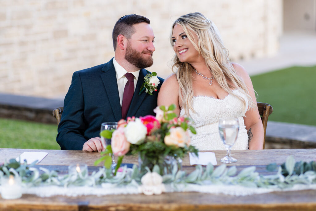 Bride and groom sitting at rustic sweetheart table smiling at each other