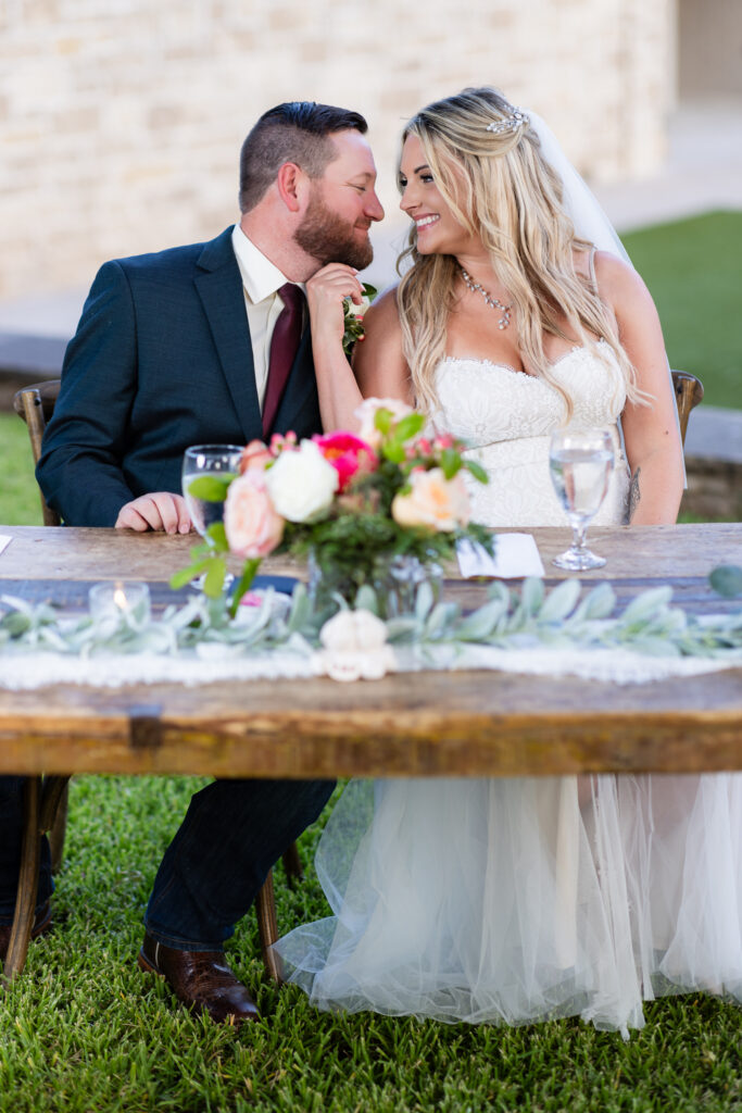 Bride and groom sitting at a rustic sweetheart table smiling closely at each other