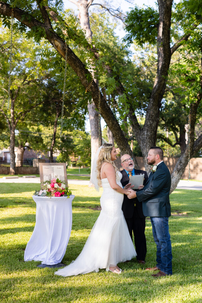 Bride and groom holding hands under lush trees during intimate elopement wedding ceremony on lawn at Hotel Lucy in Granbury Texas