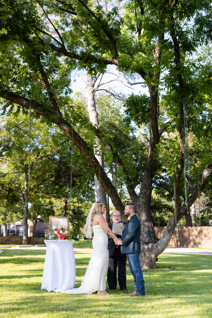 Bride and groom smiling and holding hands during intimate elopement wedding ceremony under large trees on lawn at Hotel Lucy in Granbury