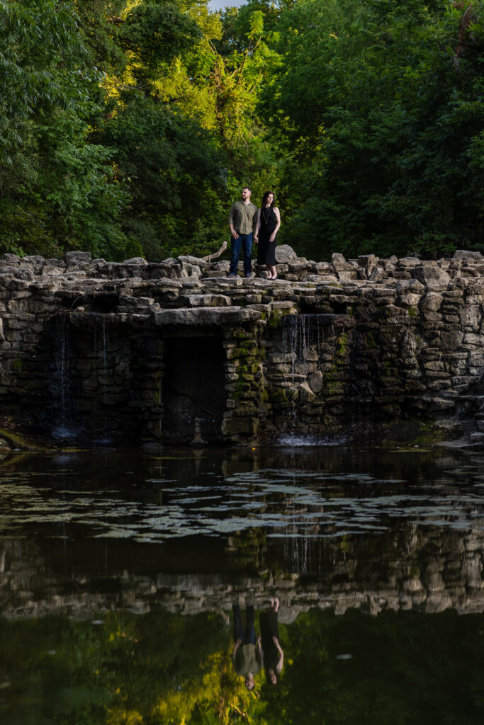 Engaged couple standing atop a stoned waterfall surrounded by lush trees during Richardson Park engagement session in Texas