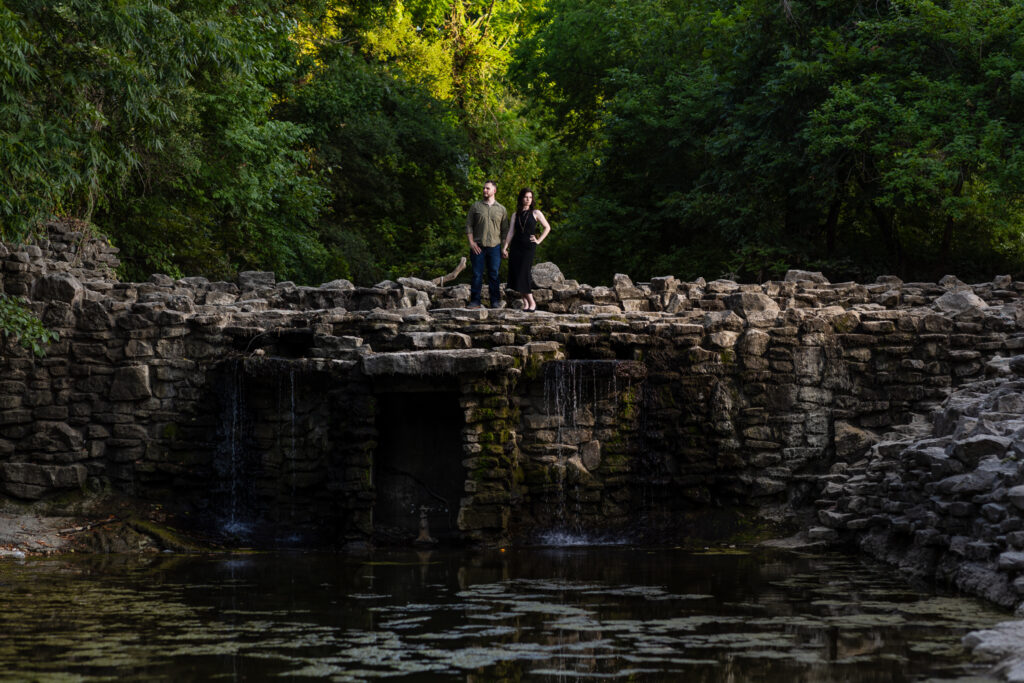 Engaged couple standing on top of a stoned waterfall surrounded by lush trees during Richardson Park engagement session in Texas