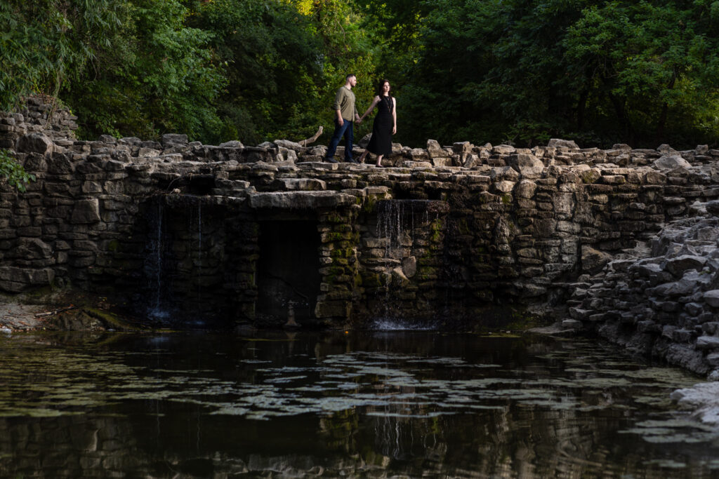 Engaged couple standing on top of a stoned waterfall surrounded by lush trees during Richardson Park engagement session in TX