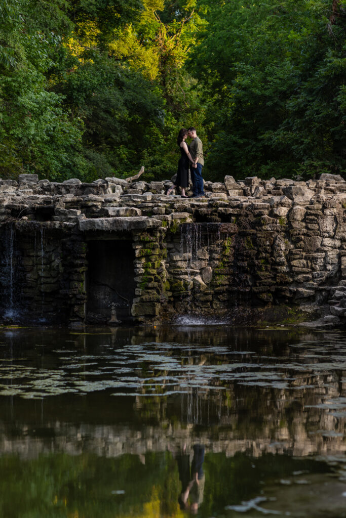 Engaged couple standing on top of a unique stoned waterfall surrounded by lush trees during Richardson Park engagement session in Texas