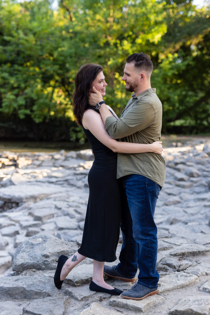 Engaged couple standing on stones with trees in the background while man cups womans face gently