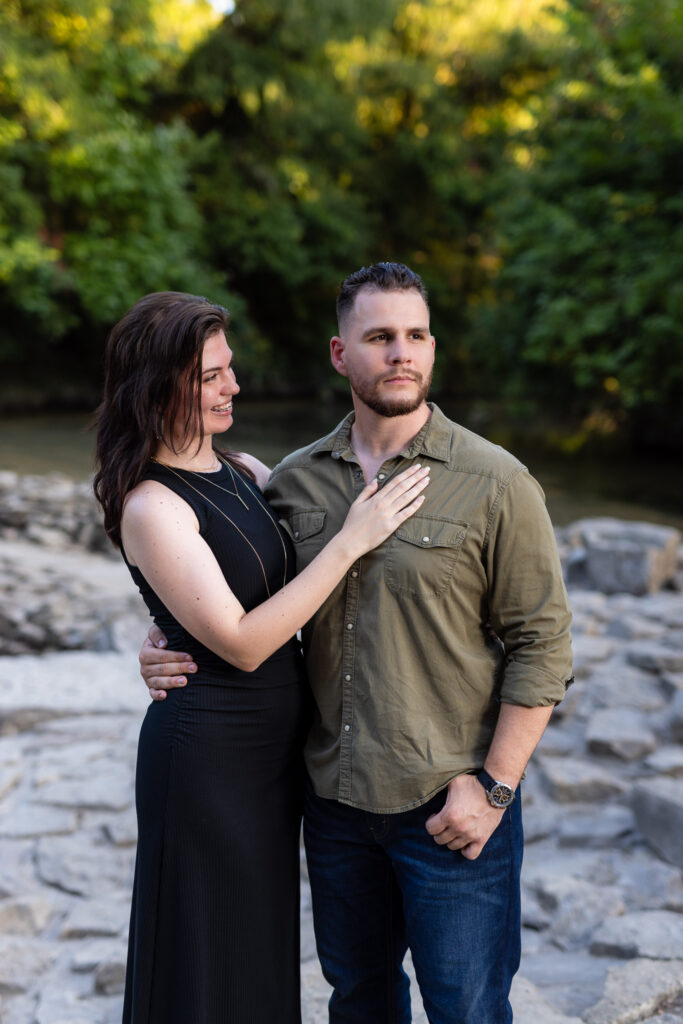 Smiling engaged woman looking at fiancé with hand on his chest surrounded by stones and trees during Richardson Park engagement session in TX